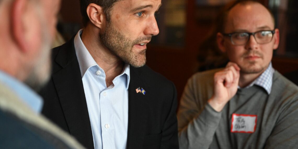 Man in a dark suit and light blue shirt speaks to colleagues during a meeting, with a bearded profile view and pins on his lapel.