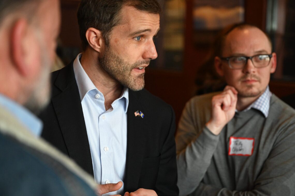 Man in a dark suit and light blue shirt speaks to colleagues during a meeting, with a bearded profile view and pins on his lapel.