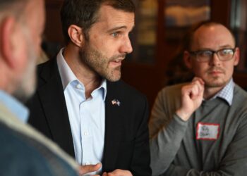 Man in a dark suit and light blue shirt speaks to colleagues during a meeting, with a bearded profile view and pins on his lapel.