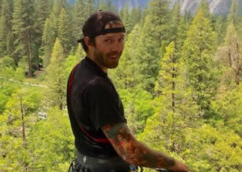 Man with a black cap backward, wearing a harness and climbing gear, standing on a platform in a pine forest with mountains in the background, looking at the camera.