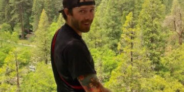 Man with a black cap backward, wearing a harness and climbing gear, standing on a platform in a pine forest with mountains in the background, looking at the camera.
