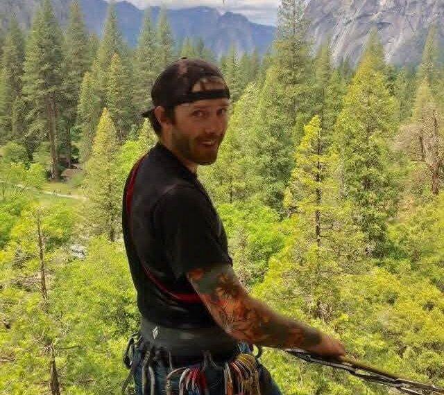 Man with a black cap backward, wearing a harness and climbing gear, standing on a platform in a pine forest with mountains in the background, looking at the camera.