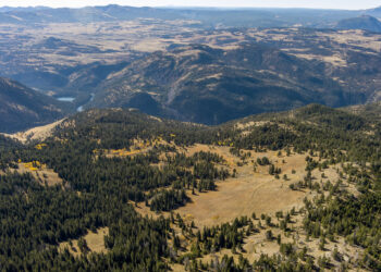 Aerial view of distant mountains, rolling hills, and pine forests with a golden grass plain and a small blue lake in the valley below.