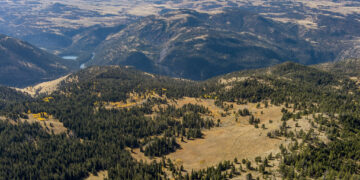 Aerial view of distant mountains, rolling hills, and pine forests with a golden grass plain and a small blue lake in the valley below.