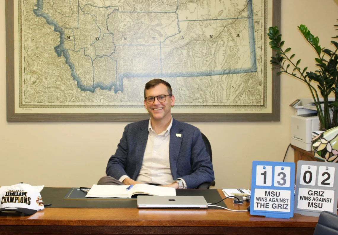 Smiling man in a blue blazer sits at a desk in an office; a large vintage map covers the wall behind him with a plant nearby.