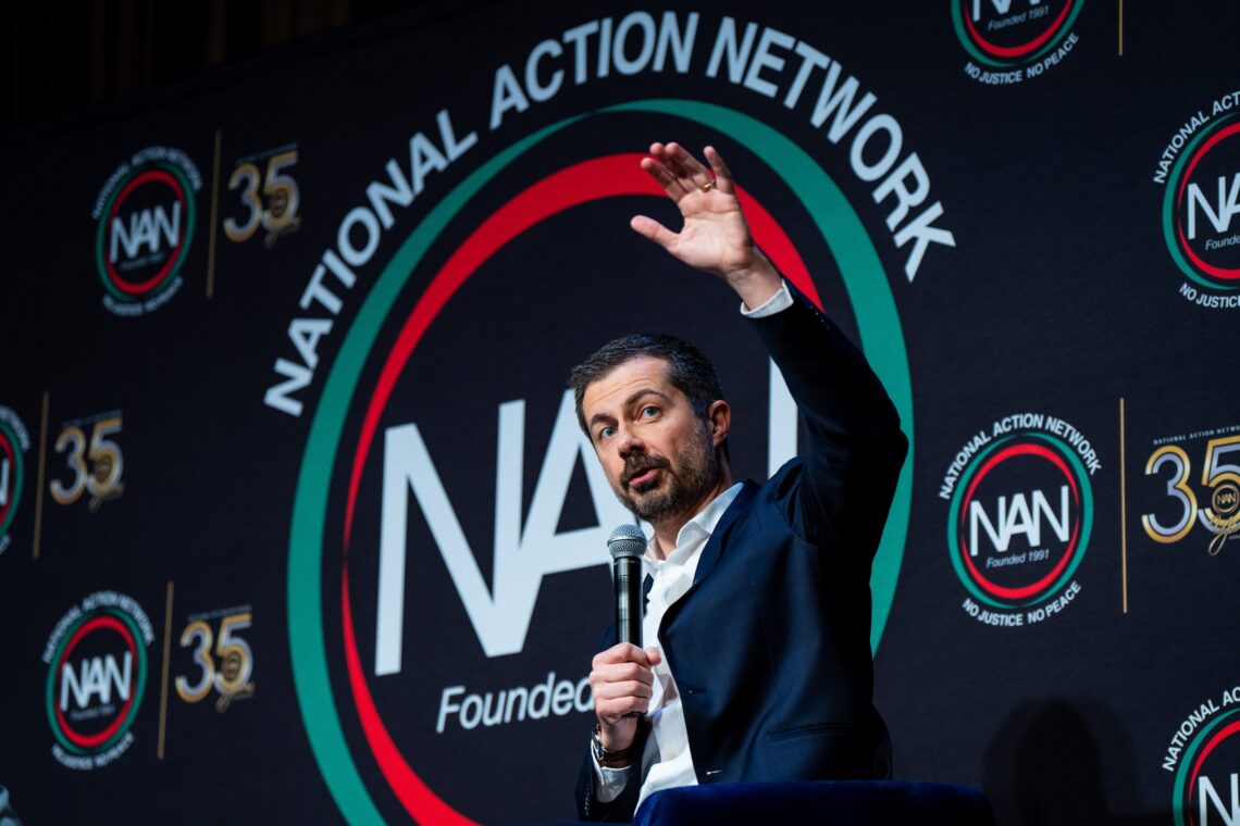 Man in a dark suit standing at a podium, raising his hand as he speaks at a National Action Network event, with NAN logos on the backdrop