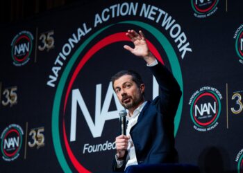 Man in a dark suit standing at a podium, raising his hand as he speaks at a National Action Network event, with NAN logos on the backdrop