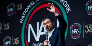 Man in a dark suit standing at a podium, raising his hand as he speaks at a National Action Network event, with NAN logos on the backdrop