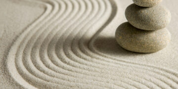 Four smooth stones stacked on sandy surface with concentric raked patterns, creating a peaceful Zen scene.
