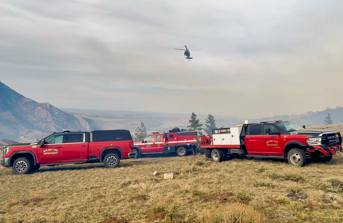 Red Big Sky Fire Department trucks parked on a grassy field with a helicopter hovering in a cloudy sky above the hills and trees in the distance.