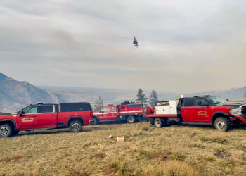 Red Big Sky Fire Department trucks parked on a grassy field with a helicopter hovering in a cloudy sky above the hills and trees in the distance.