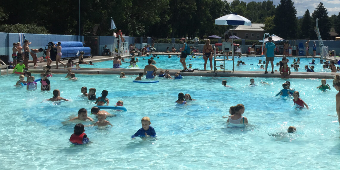 Crowded outdoor public pool with kids swimming, lifeguards on duty, and shade umbrellas along the deck.