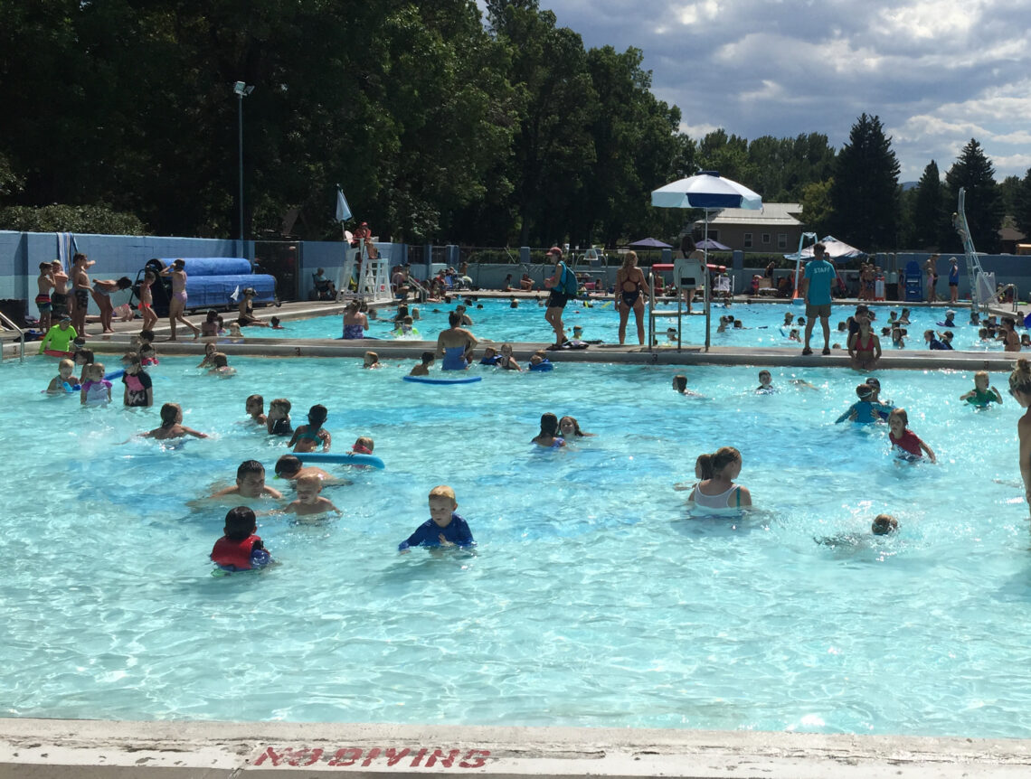 Crowded outdoor public pool with kids swimming, lifeguards on duty, and shade umbrellas along the deck.