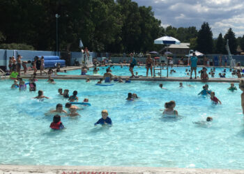 Crowded outdoor public pool with kids swimming, lifeguards on duty, and shade umbrellas along the deck.
