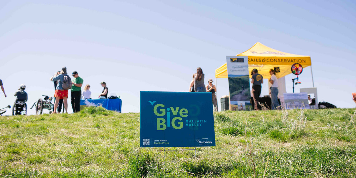Blue GiveBIG Gallatin Valley sign in a grassy field at an outdoor event with tents and people in the background.