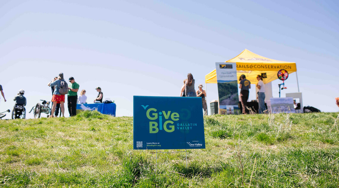 Blue GiveBIG Gallatin Valley sign in a grassy field at an outdoor event with tents and people in the background.