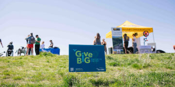 Blue GiveBIG Gallatin Valley sign in a grassy field at an outdoor event with tents and people in the background.