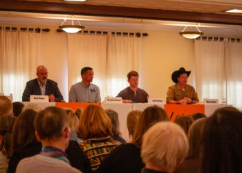 Panel of four men seated at a long table on a stage, facing an audience; nameplates read Ryan Busse, Russell Cleveland, Sam Forstagn, and Matt Rains in front of them.