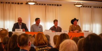 Panel of four men seated at a long table on a stage, facing an audience; nameplates read Ryan Busse, Russell Cleveland, Sam Forstagn, and Matt Rains in front of them.