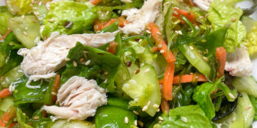 Bowl of mixed green salad with shredded chicken, cucumber, carrot, and sesame seeds.