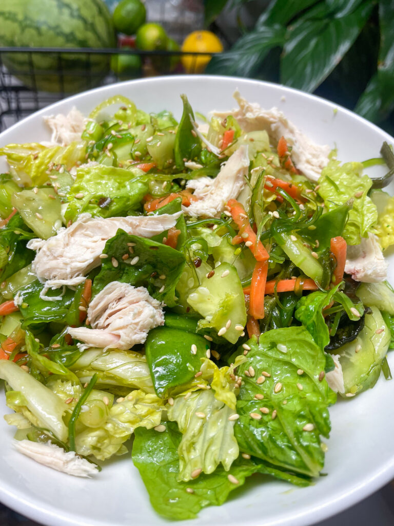 Bowl of mixed green salad with shredded chicken, cucumber, carrot, and sesame seeds.