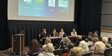 Panel discussion with four speakers seated at a long table on a stage; projection screen shows logos and the names Anne, Barbara, Amy, Derek.