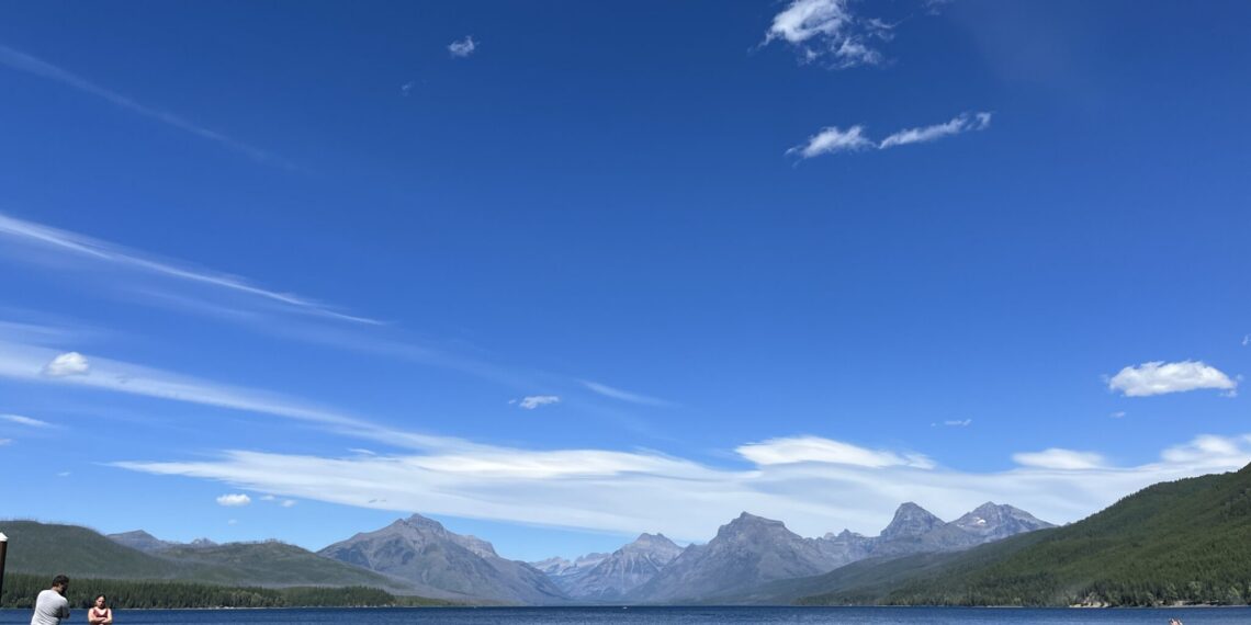 People stand on a wooden dock at a calm mountain lake, with clear blue sky and rugged peaks in the distance.