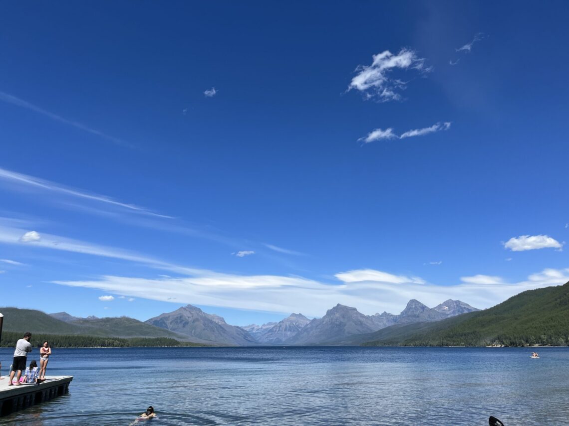 People stand on a wooden dock at a calm mountain lake, with clear blue sky and rugged peaks in the distance.