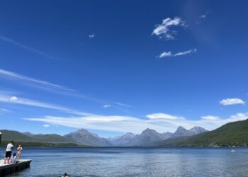 People stand on a wooden dock at a calm mountain lake, with clear blue sky and rugged peaks in the distance.