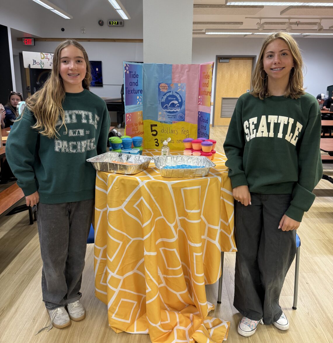 Two teenage girls in Seattle sweatshirts stand at a table with a bright orange and yellow project board titled 'Tide and Texture' in a classroom setting.