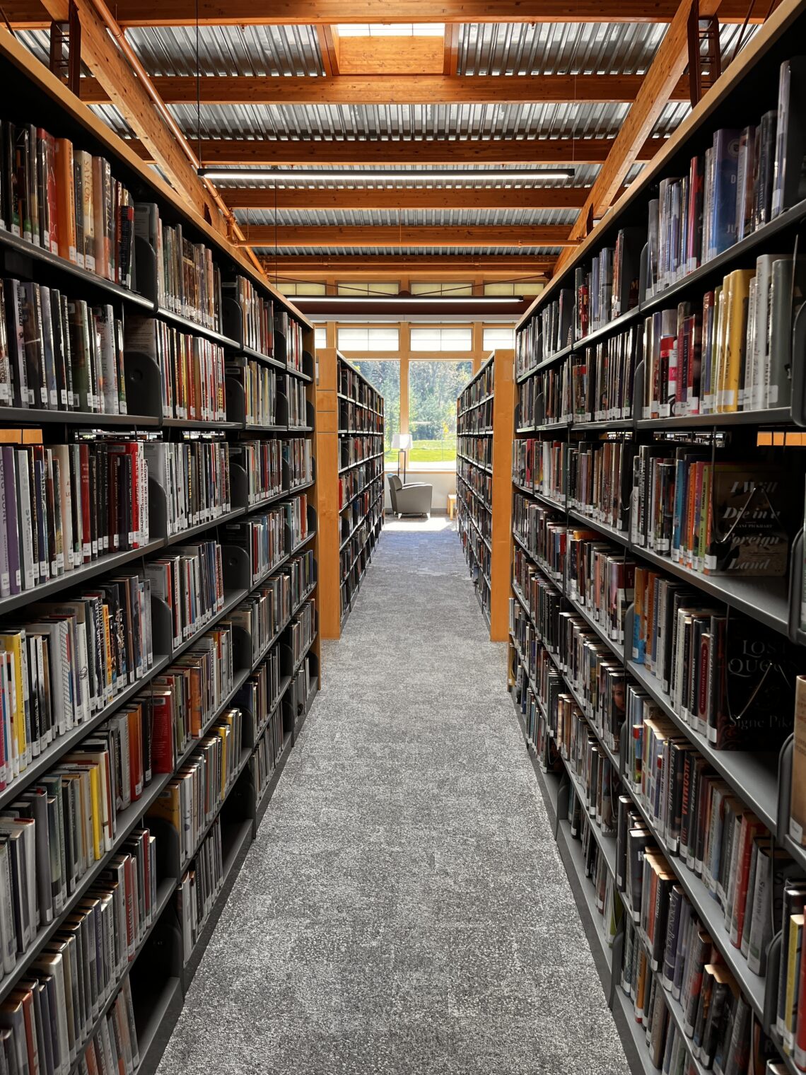 Long library aisle between tall shelves filled with books, leading to a window at the far end with seating nearby.