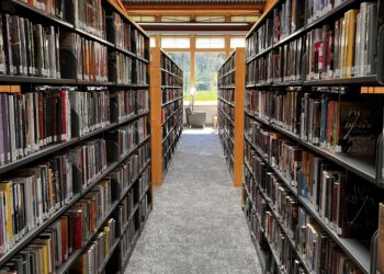 Long library aisle between tall shelves filled with books, leading to a window at the far end with seating nearby.
