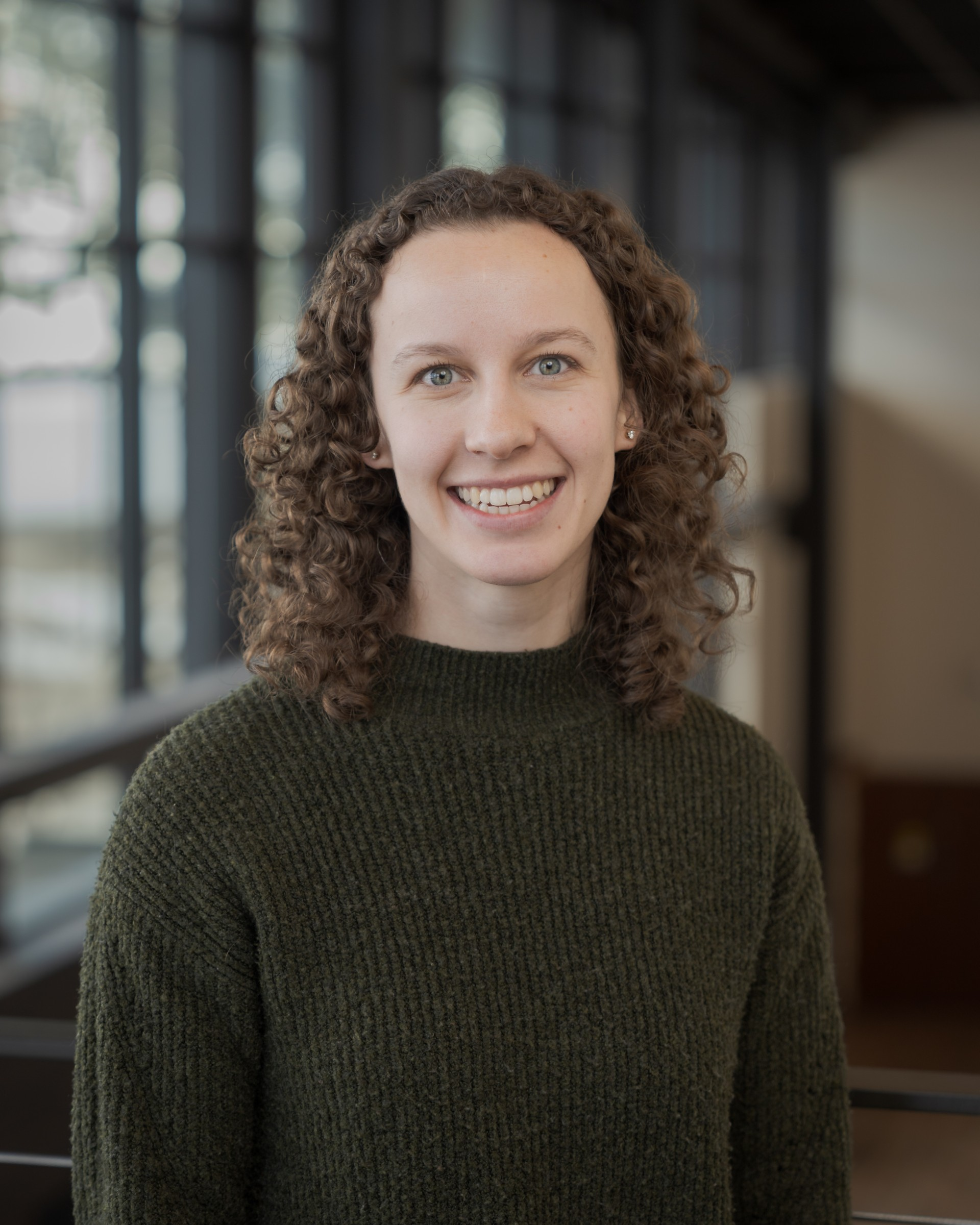 Close-up portrait of a woman with curly brown hair and a dark green knit sweater smiling at the camera in a modern indoor space with blurred background.