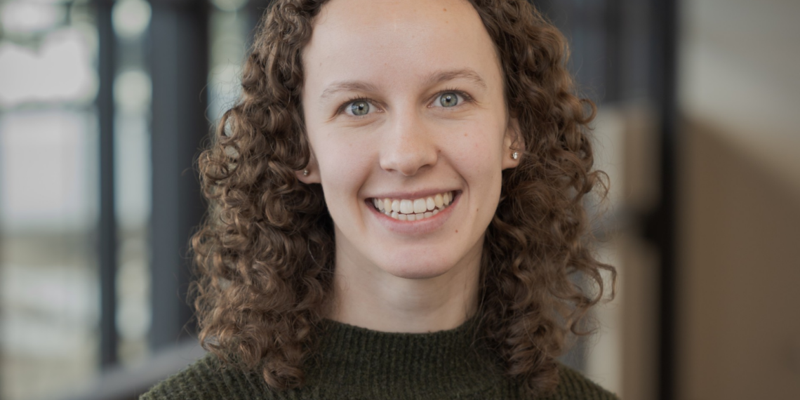 Smiling woman with curly brown hair wearing a dark green knit sweater, indoors in a softly blurred background.