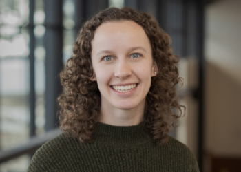 Smiling woman with curly brown hair wearing a dark green knit sweater, indoors in a softly blurred background.