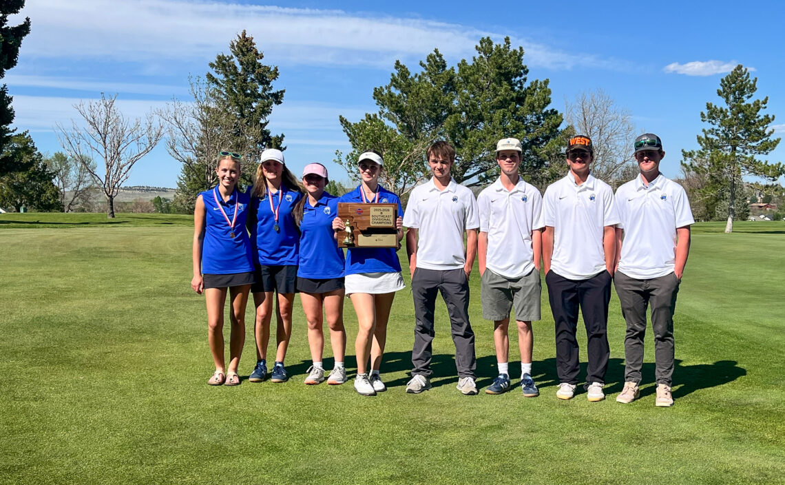Eight young golfers pose for a team photo on a sunny golf course, wearing blue and white shirts with medals and a trophy plaque in the center.