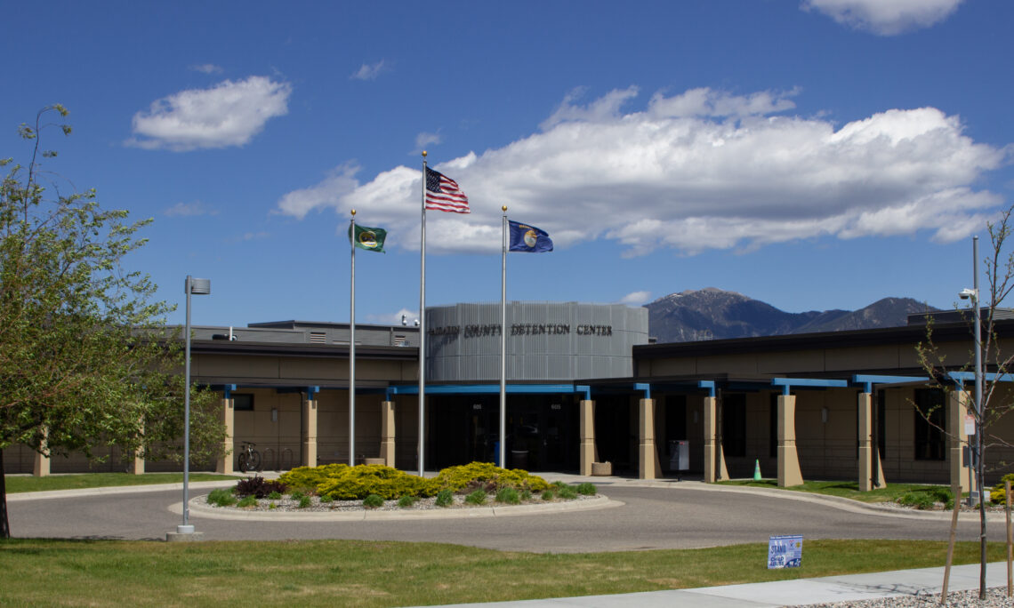 Front view of a county detention center with three flagpoles—US flag and two state flags—flying against a blue sky with mountains in the distance.