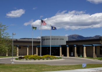 Front view of a county detention center with three flagpoles—US flag and two state flags—flying against a blue sky with mountains in the distance.