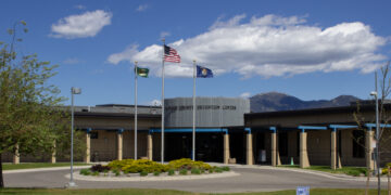 Front view of a county detention center with three flagpoles—US flag and two state flags—flying against a blue sky with mountains in the distance.