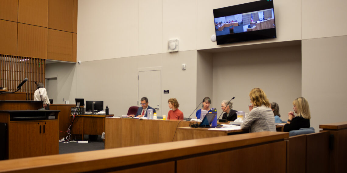 Panel of people seated at a long wood conference table in a formal meeting room with a wall-mounted screen showing a courtroom scene.