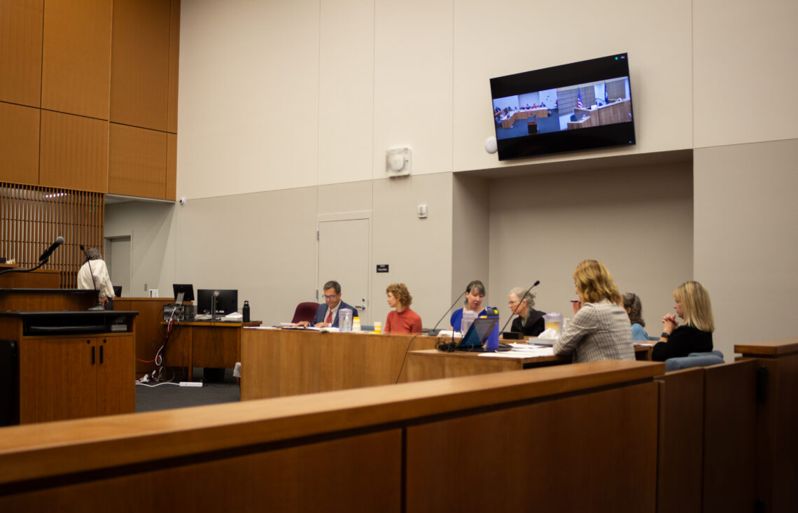 Panel of people seated at a long wood conference table in a formal meeting room with a wall-mounted screen showing a courtroom scene.