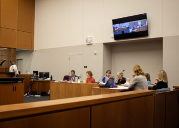 Panel of people seated at a long wood conference table in a formal meeting room with a wall-mounted screen showing a courtroom scene.