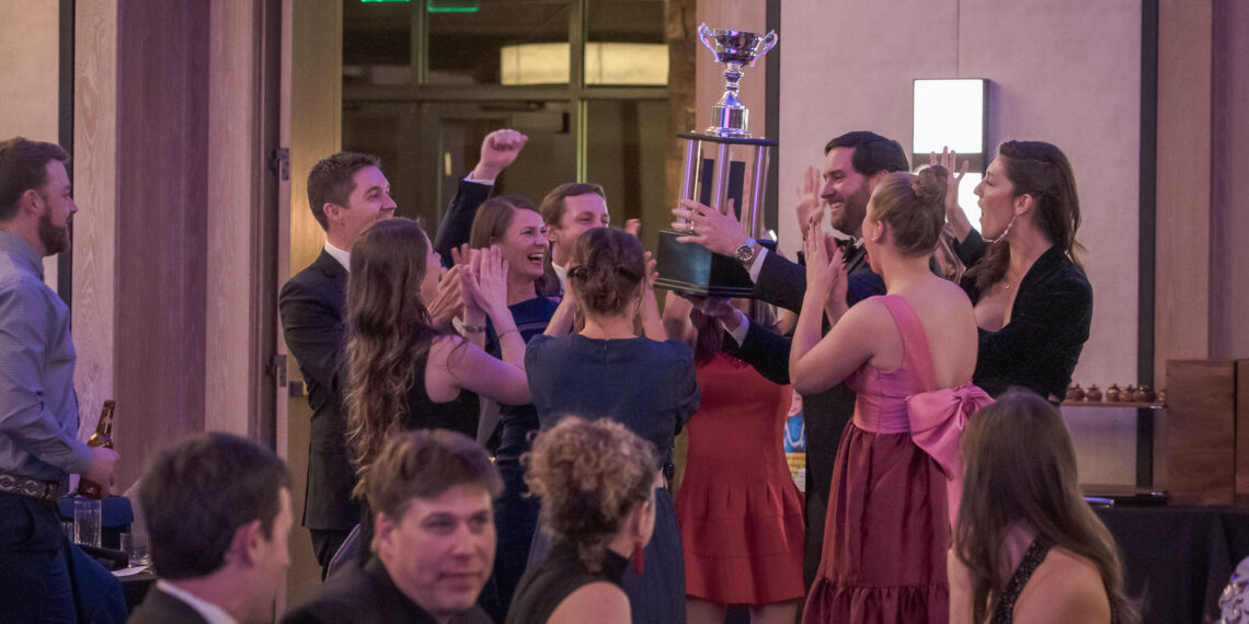 Group of formally dressed people cheering as they lift a tall trophy in a celebration indoors, pink dresses and suits visible in the crowd.
