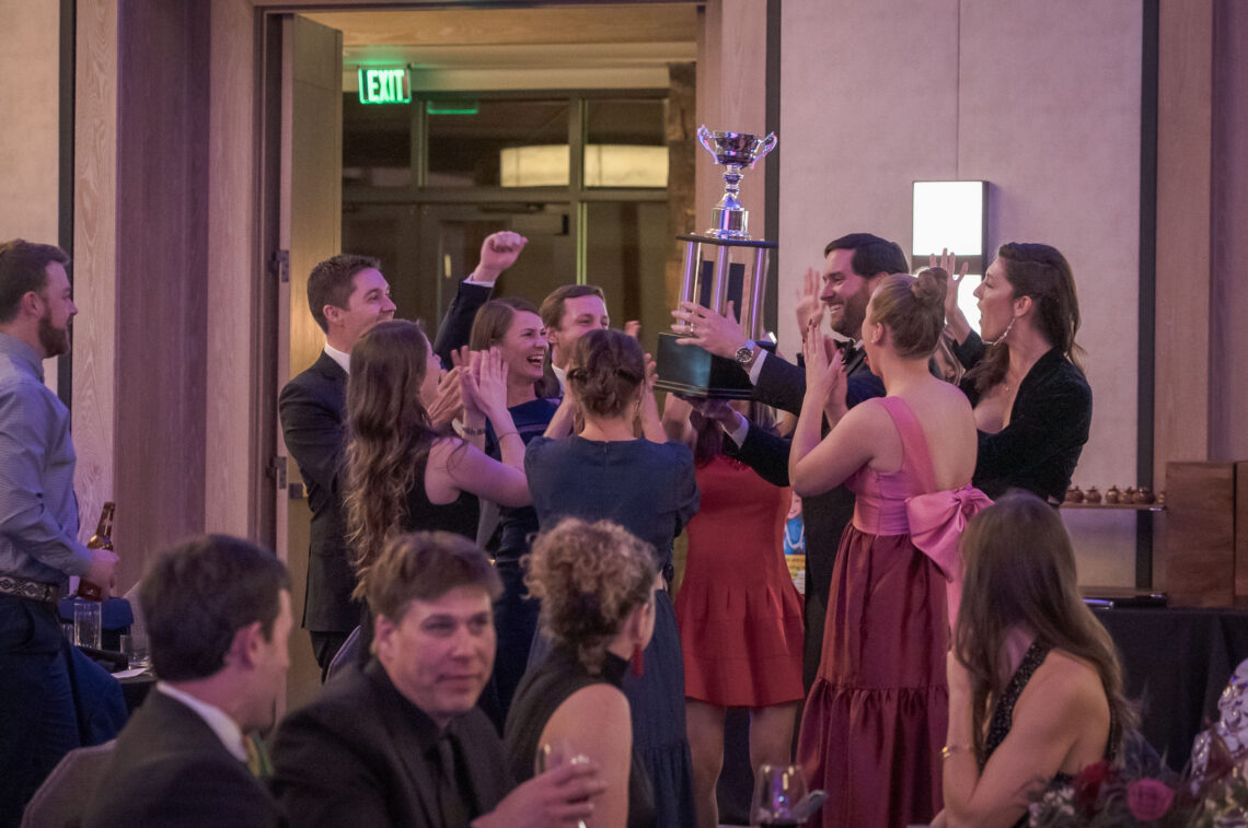 Group of formally dressed people cheering as they lift a tall trophy in a celebration indoors, pink dresses and suits visible in the crowd.