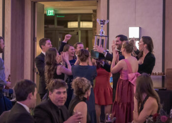 Group of formally dressed people cheering as they lift a tall trophy in a celebration indoors, pink dresses and suits visible in the crowd.