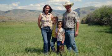 Family of four posing in a grassy field with rolling hills in the background, sunny day.