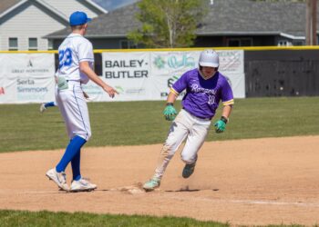 Two baseball players near a base: one in a white uniform with blue cap approaching a bag, the other in purple Rangers jersey sliding toward home plate.