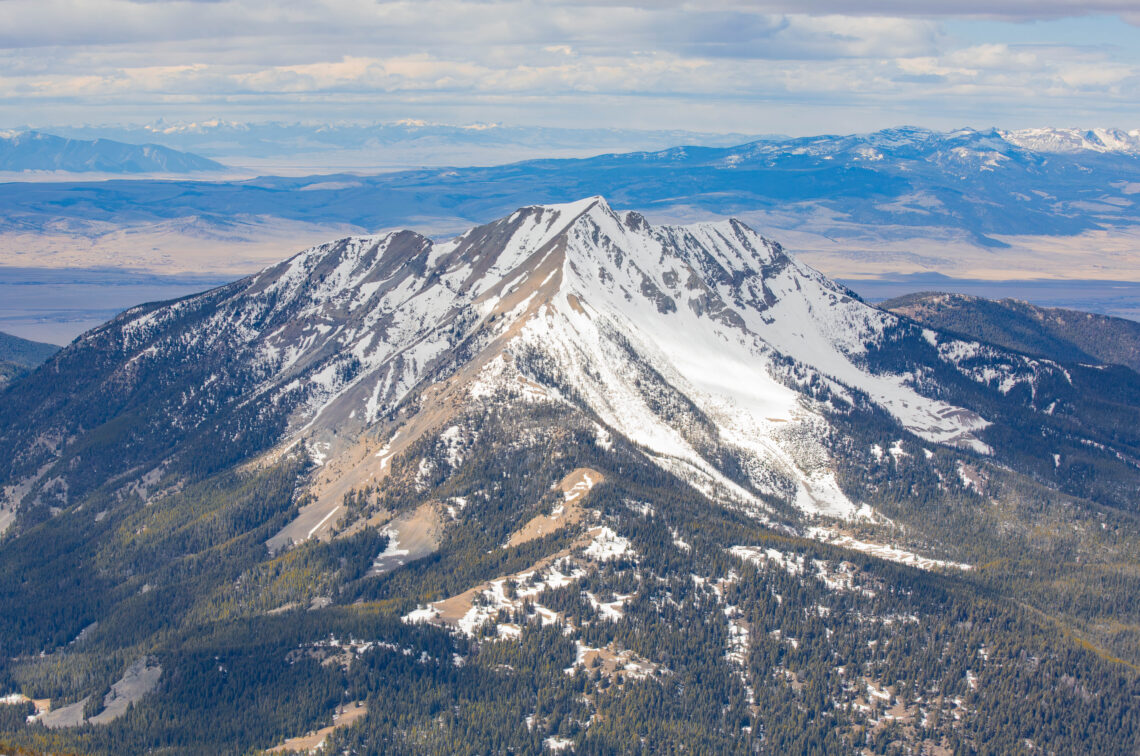 Snow-covered mountain peak in the foreground with forested slopes and distant blue ranges under a cloudy sky.