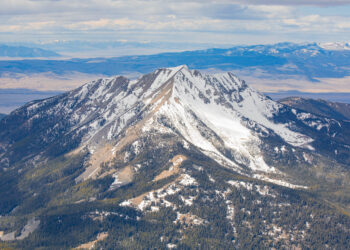 Snow-covered mountain peak in the foreground with forested slopes and distant blue ranges under a cloudy sky.