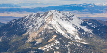 Snow-covered mountain peak in the foreground with forested slopes and distant blue ranges under a cloudy sky.